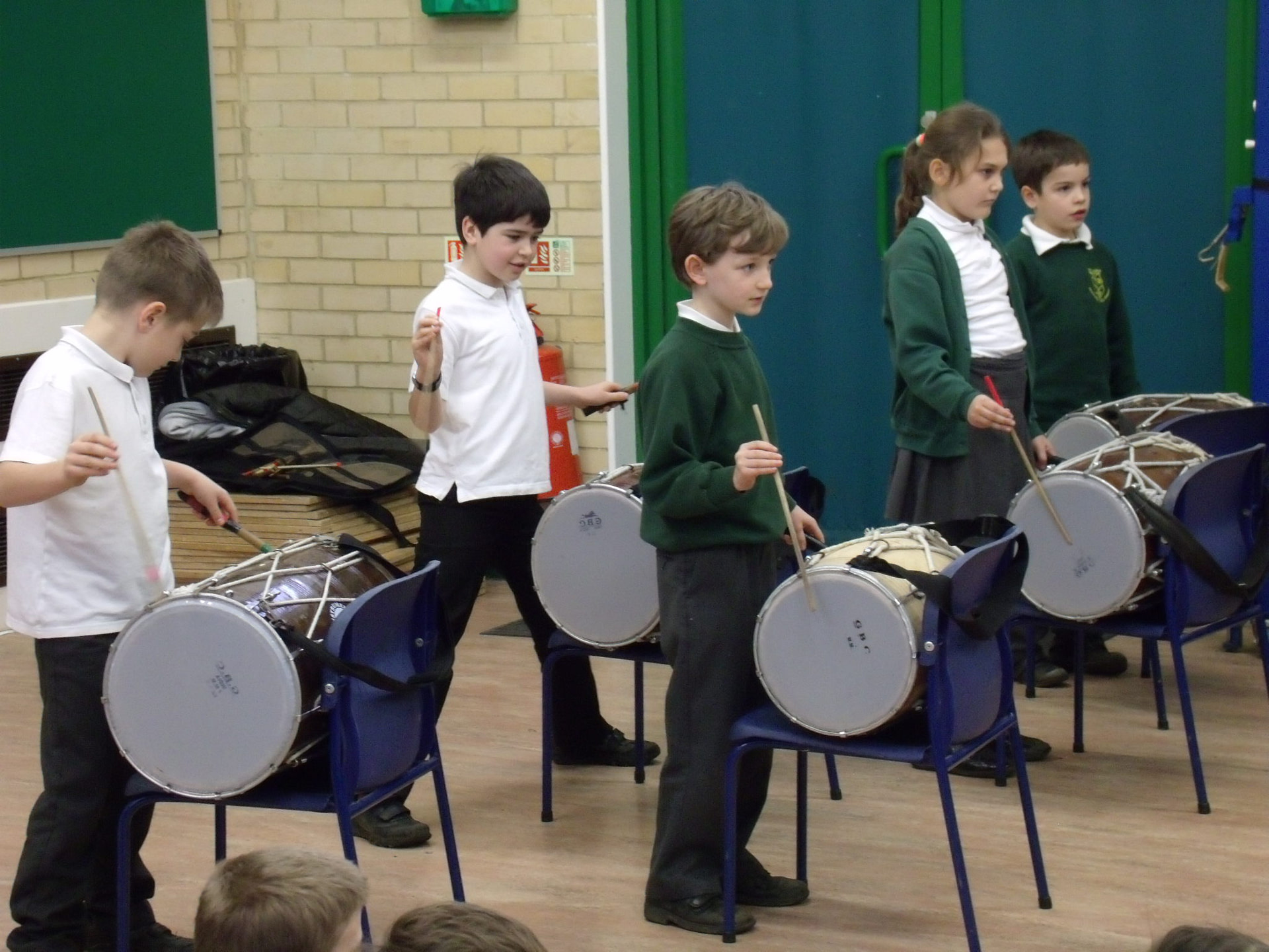 School children playing Indian drums in school hall on chairs
