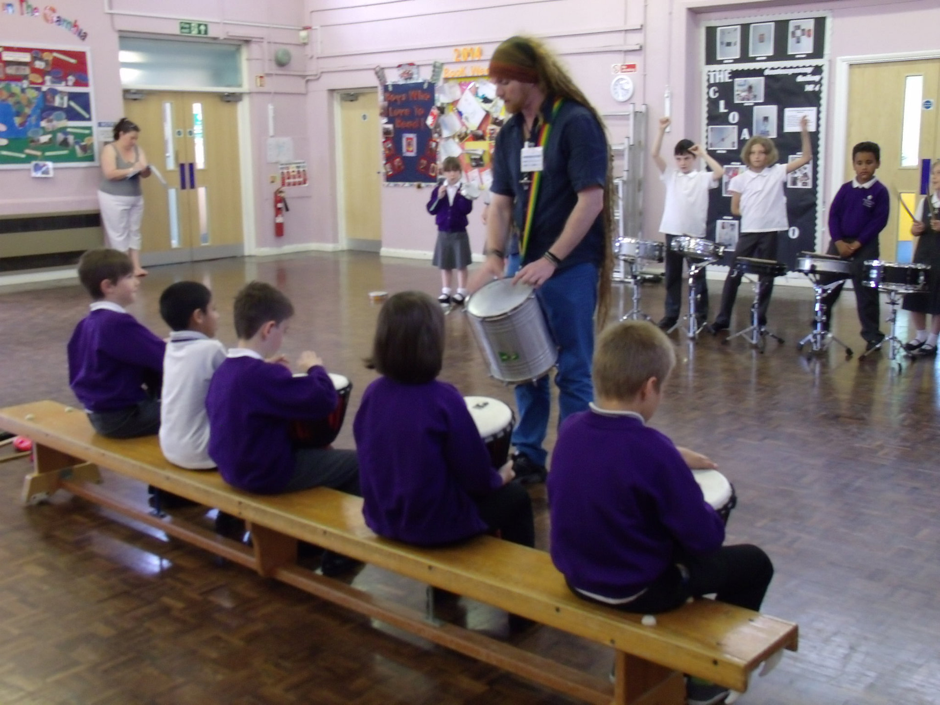 Teacher demonstrating to school children sitting on a bench how to play Samba drums